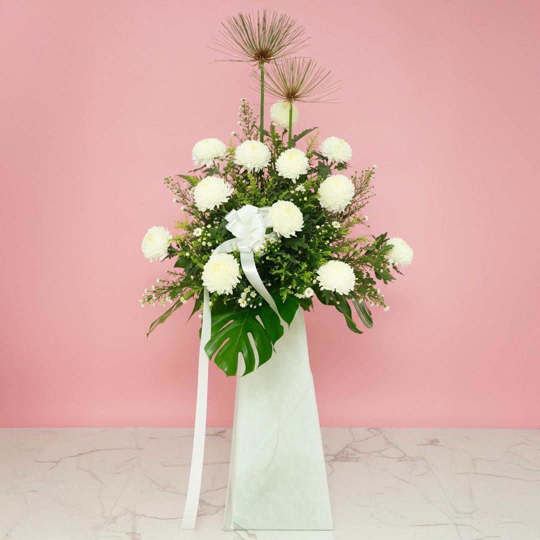 God Is With You A white stand containing funeral flowers filled with delicate white flowers and green leaves.