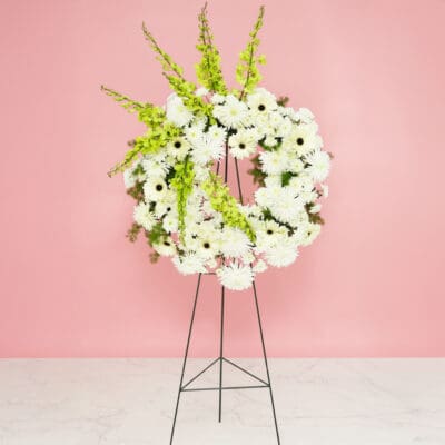 A white floral wreath adorned with green and white flowers, displayed elegantly on a stand.