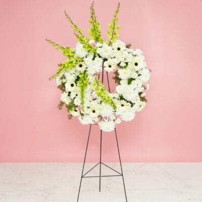 A white floral wreath adorned with green and white flowers, displayed elegantly on a stand.