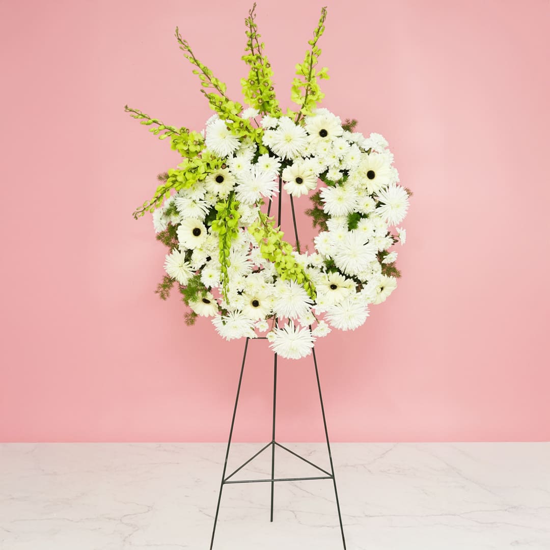 Rest In Peace A white floral wreath adorned with green and white flowers, displayed elegantly on a stand.