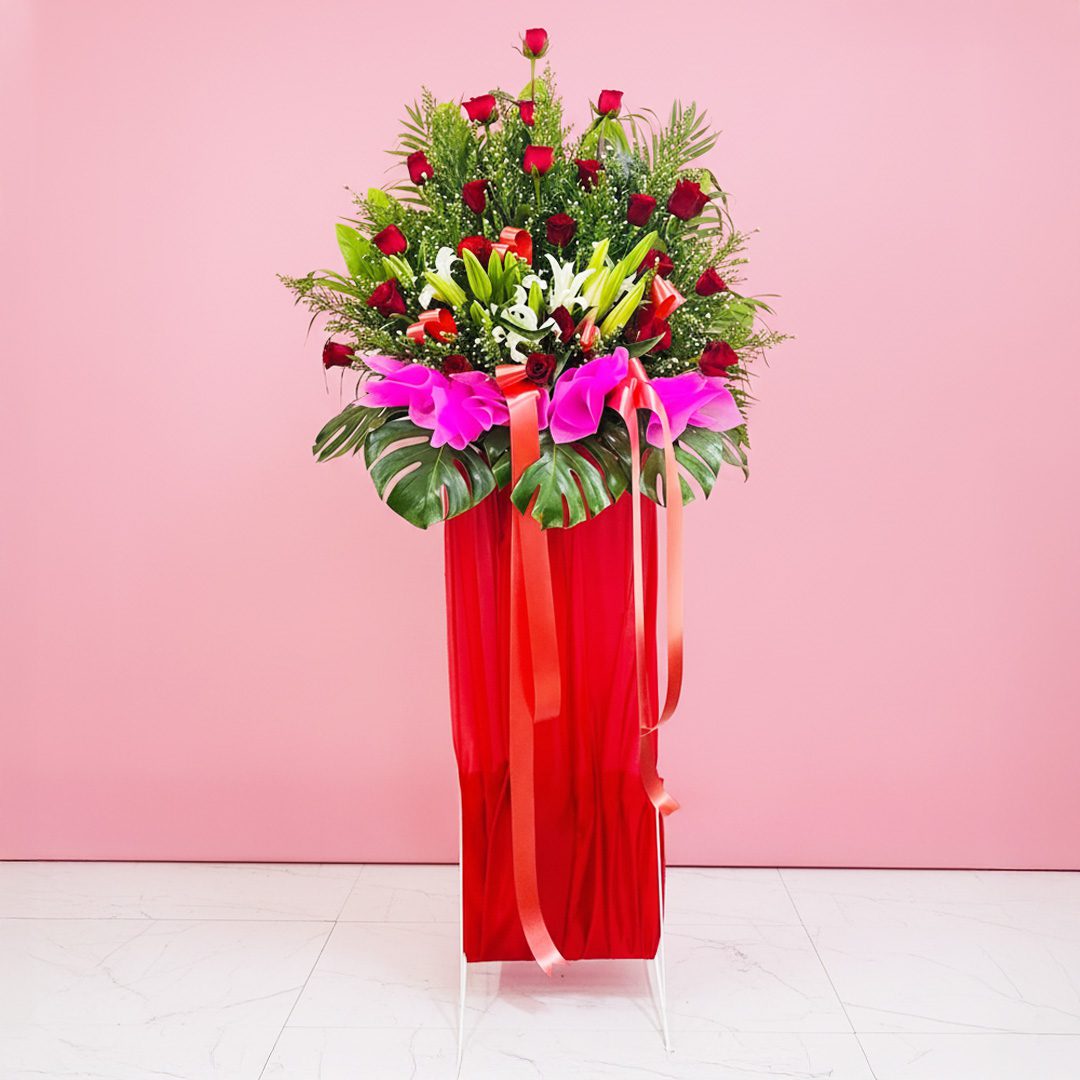 Flower Fusion Greeting Hub A red Grand Opening Stand filled with assorted flowers, including roses and lilies, adorned with a ribbon against a pink background.