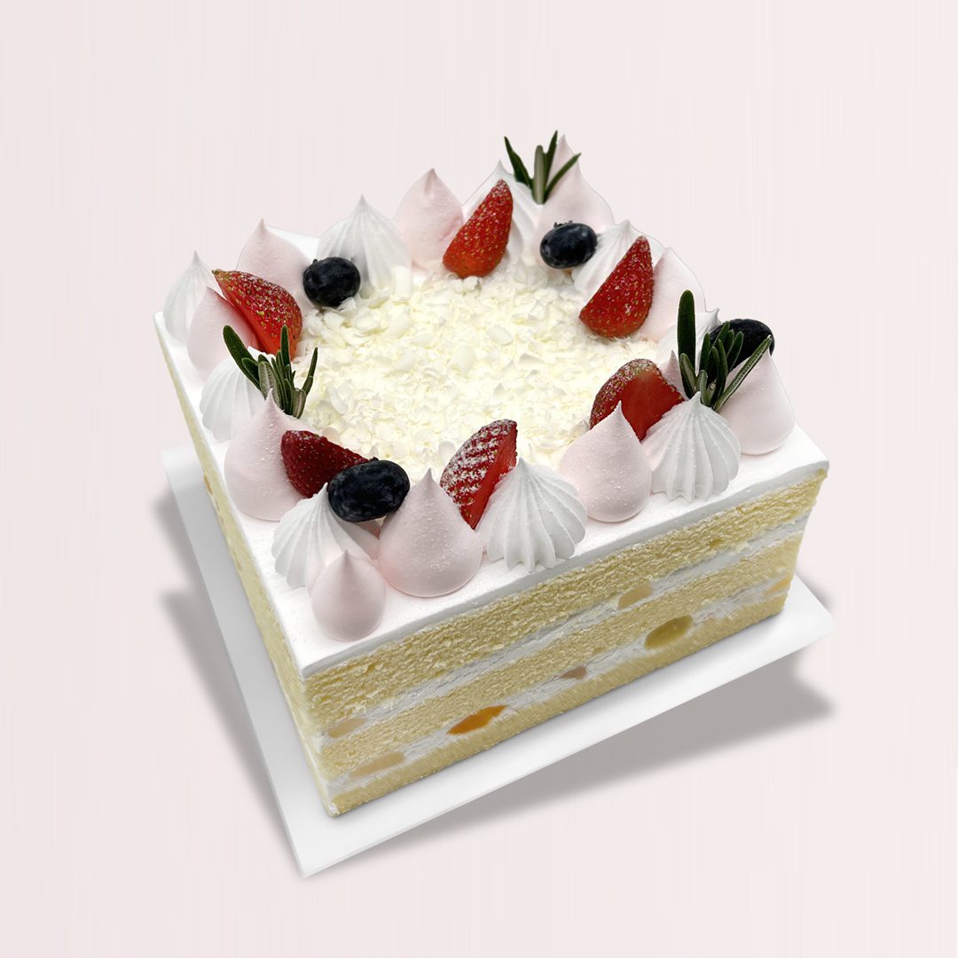 The image shows a square-shaped cake with a white background. The cake is layered with sponge cake, cream, and fruit. The top of the cake is decorated with white chocolate shavings, strawberries, blueberries, and meringue kisses.
