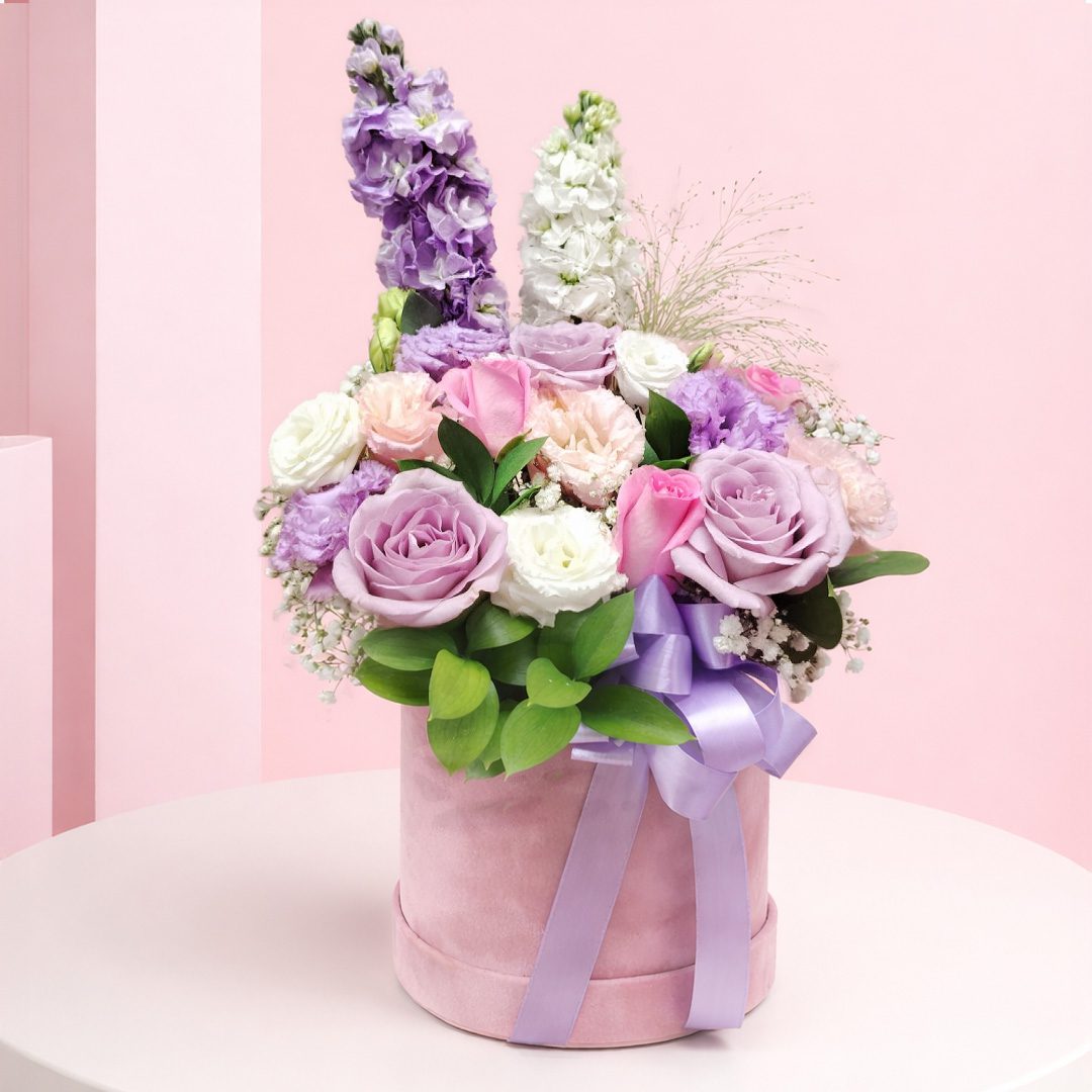 01 Lavender rose hatbox flower arrangement with pink roses, white flowers, greenery, and ribbon on a wooden table.