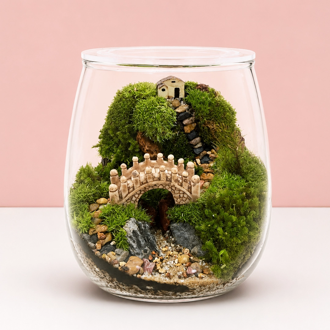 enchanted-bridge-glass-bowl-terrarium Enchanted Bridge Glass Bowl Terrarium featuring a miniature bridge, lush green moss, and a tiny cottage landscape arranged inside a decorative glass terrarium.