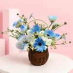 Flower basket featuring 5 blue gerberas, 1 lisianthus stem, chamomile, white wax flowers, seasonal fillers, and lush green foliage.