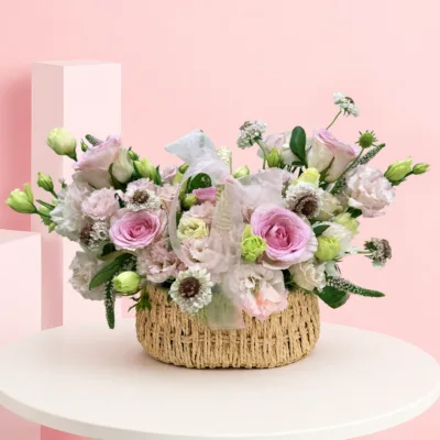 A handcrafted floral basket featuring pink roses, lisianthus, veronica, scabiosa blooms, and vibrant foliage in a naturally flowing arrangement.