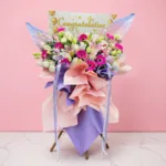 A congratulations flower stand with pink gerberas, pink lilies, white blooms, and fresh green foliage.