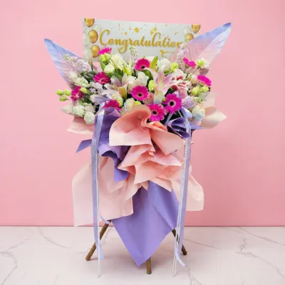A congratulations flower stand with pink gerberas, pink lilies, white blooms, and fresh green foliage.