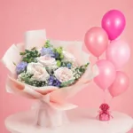 5 light pink roses, 2 purple lisianthus arranged with baby’s breath, mixed greens, and 5 colourful balloons