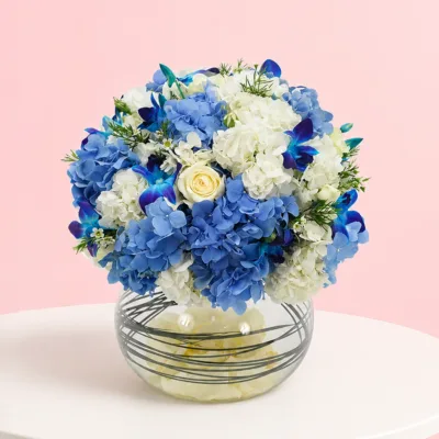 A floral arrangement featuring 3 blue hydrangeas, 1 white hydrangea, 4 blue orchids, 4 roses, and 5 wax flowers with greenery.