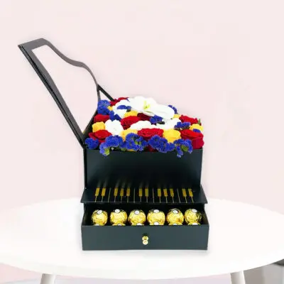 A black piano gift box surrounded by colorful flowers and an assortment of chocolates on a wooden table.