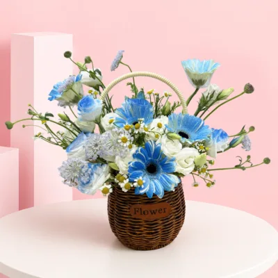 Flower basket featuring 5 blue gerberas, 1 lisianthus stem, chamomile, white wax flowers, seasonal fillers, and lush green foliage.
