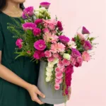 Black cylindrical flower box filled with purple toned fresh gerberas, spray roses, and seasonal foliage.
